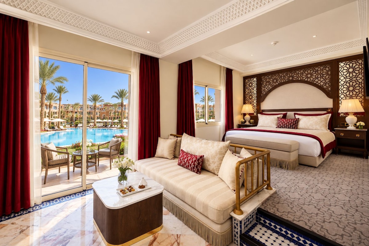 A hotel room with a bed, a sofa, a dining table, and a view of a swimming pool.