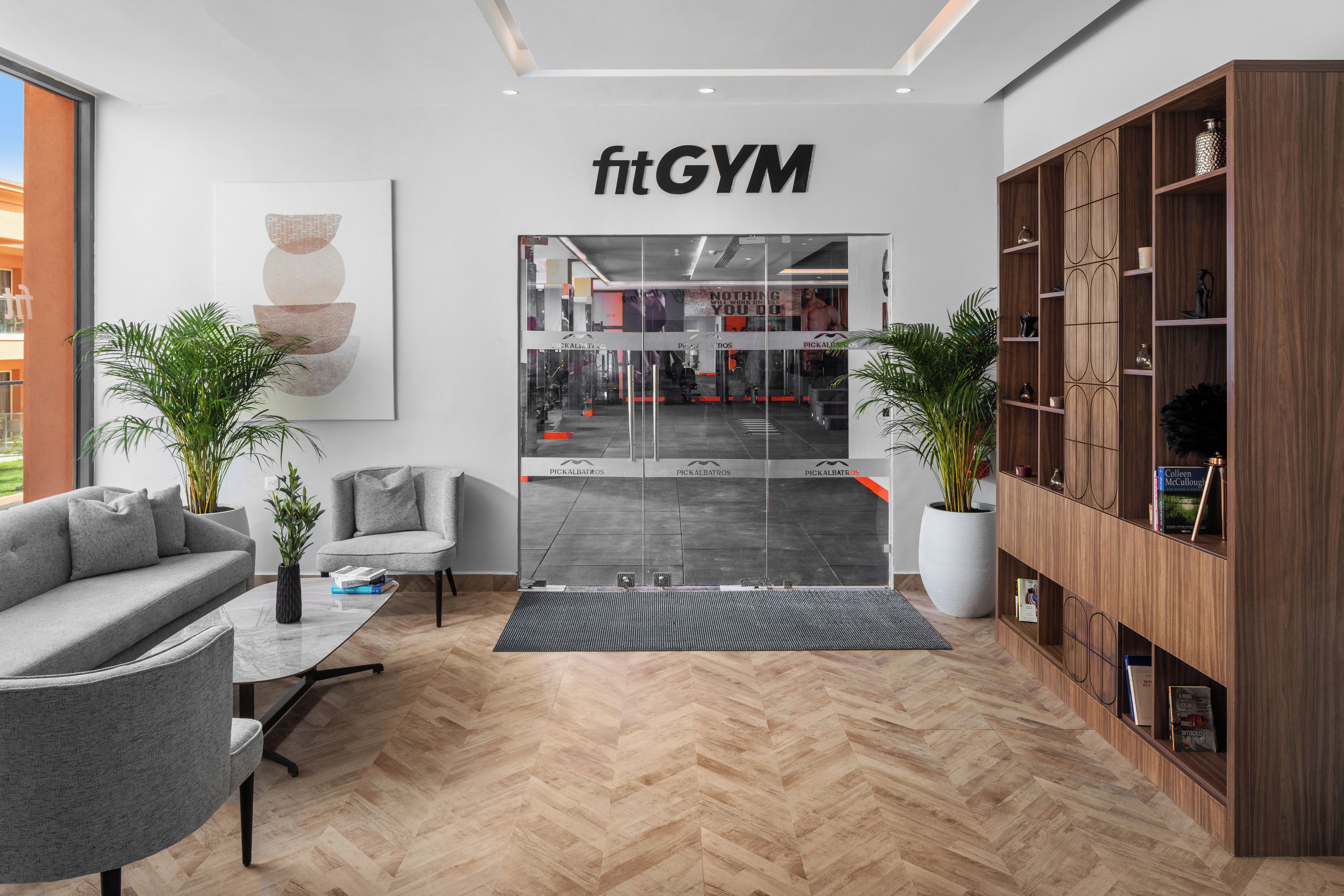 A modern gym interior with a reception area, grey sofas, wooden shelving, and a herringbone floor pattern.