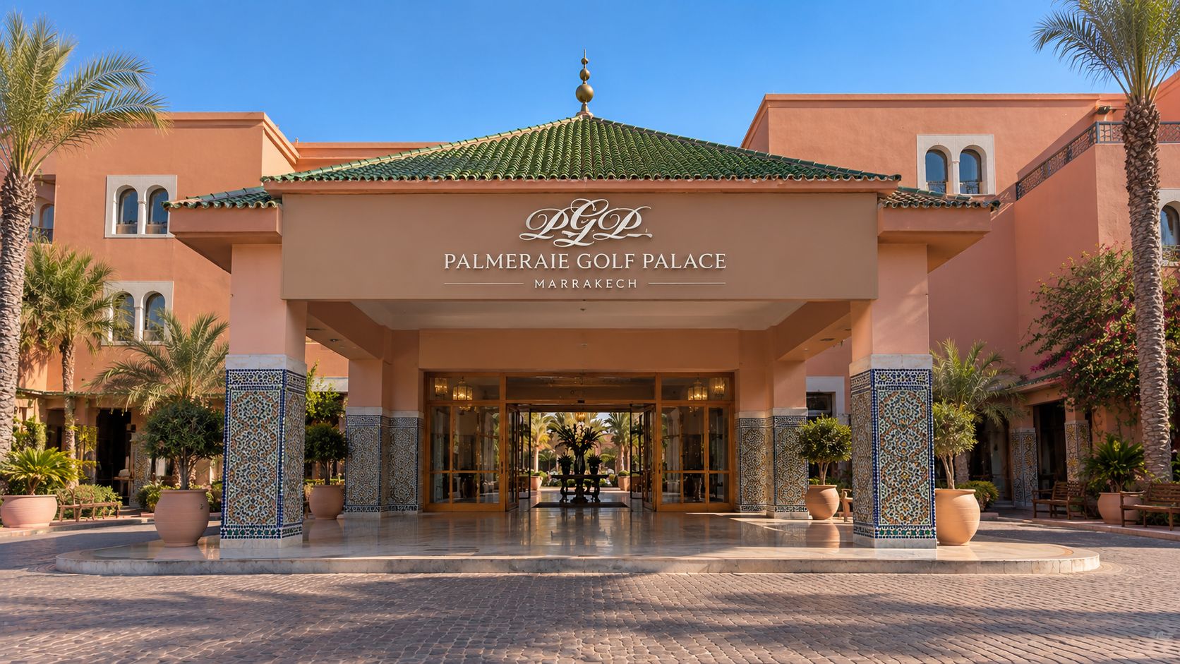 Der Eingang des Palmiera Golf Palace mit einer gefliesten Fassade, Palmen und blauem Himmel.