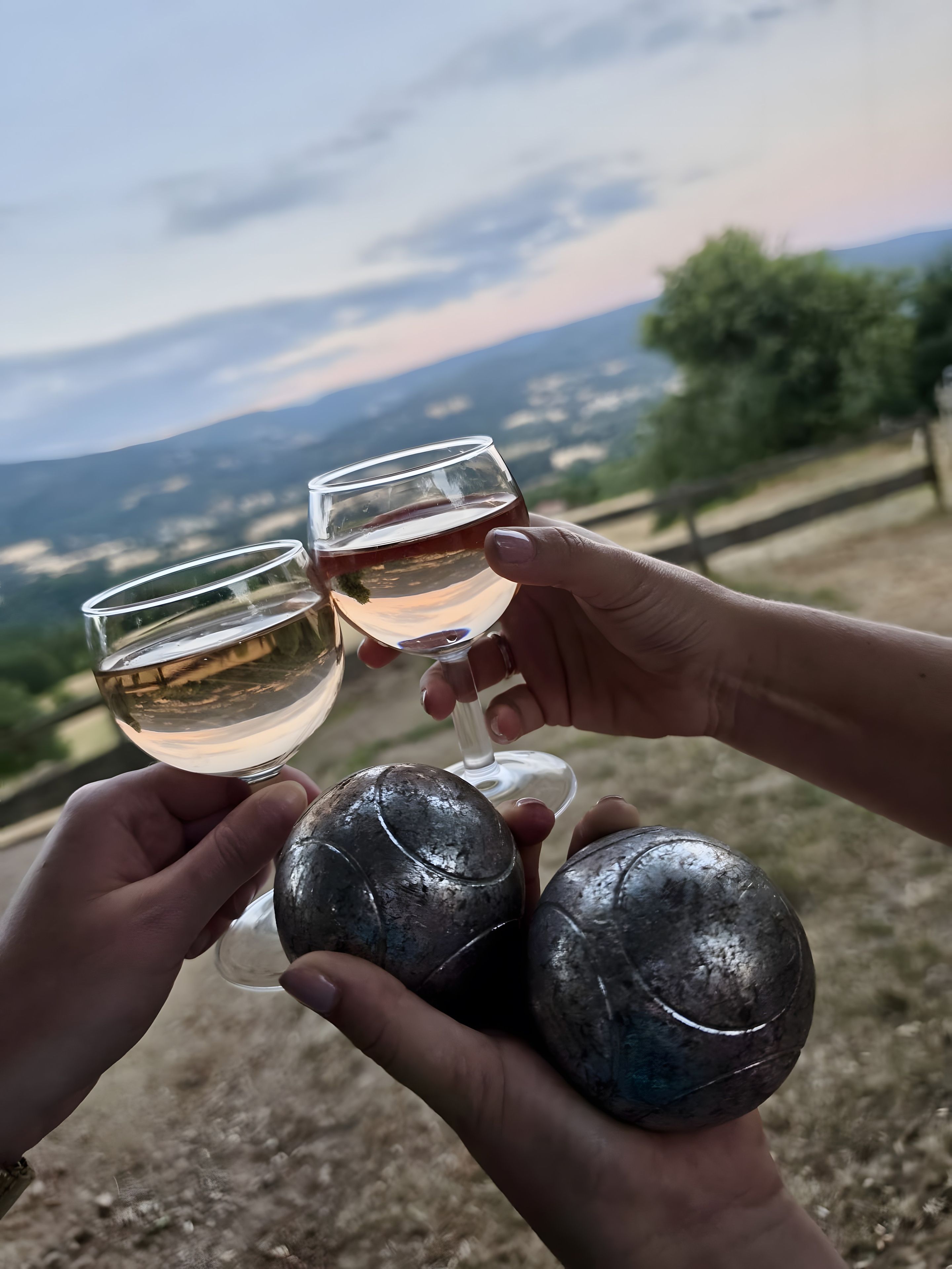 A person holding a wine glass with a clear liquid, another glass with a red liquid, and a third glass with a clear liquid, with a scenic view in the background.