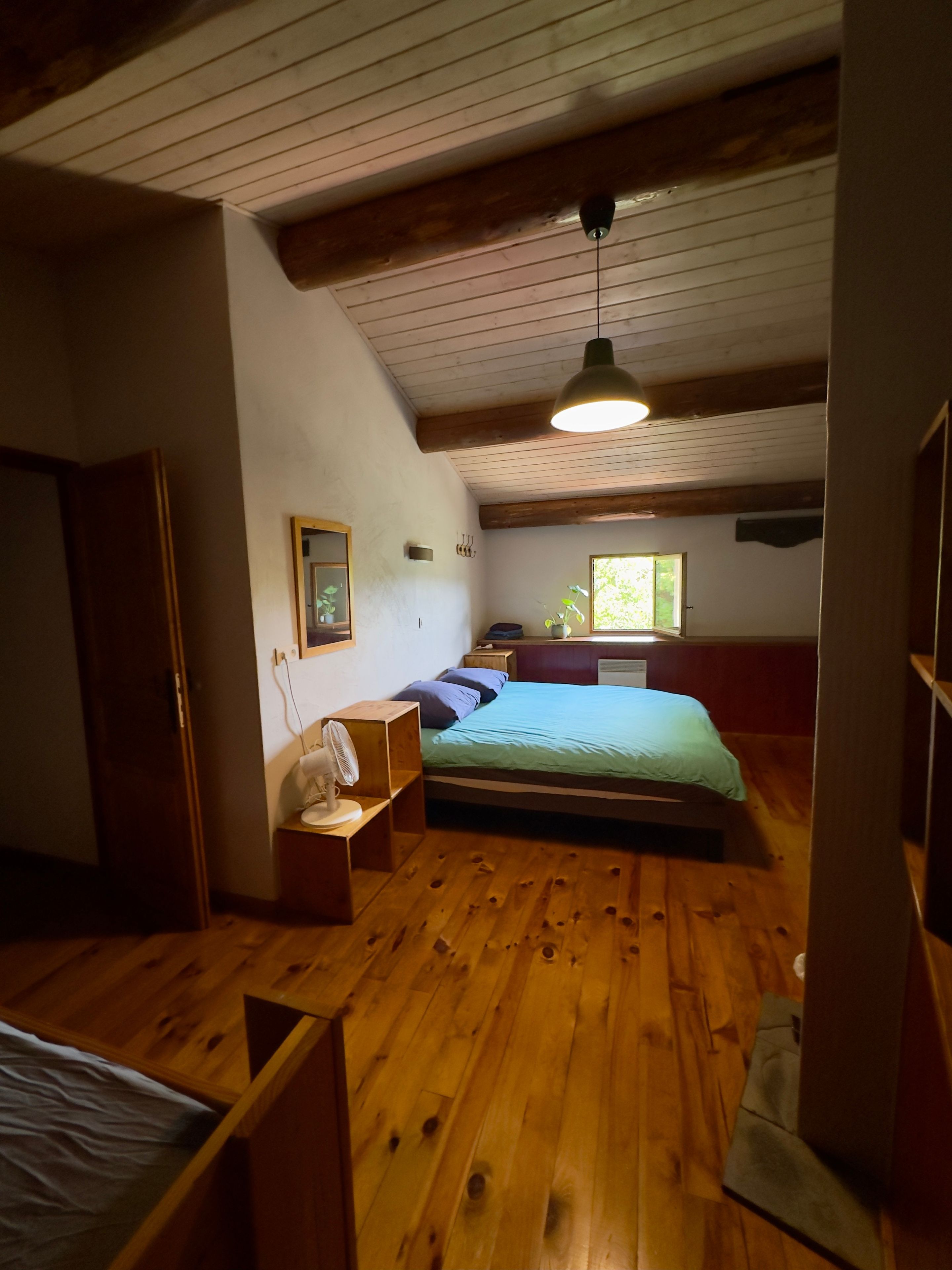 Una habitación con piso de madera, una cama con colchón azul, una mesita de noche con lámpara, una ventana que da a zonas verdes y un techo de madera.