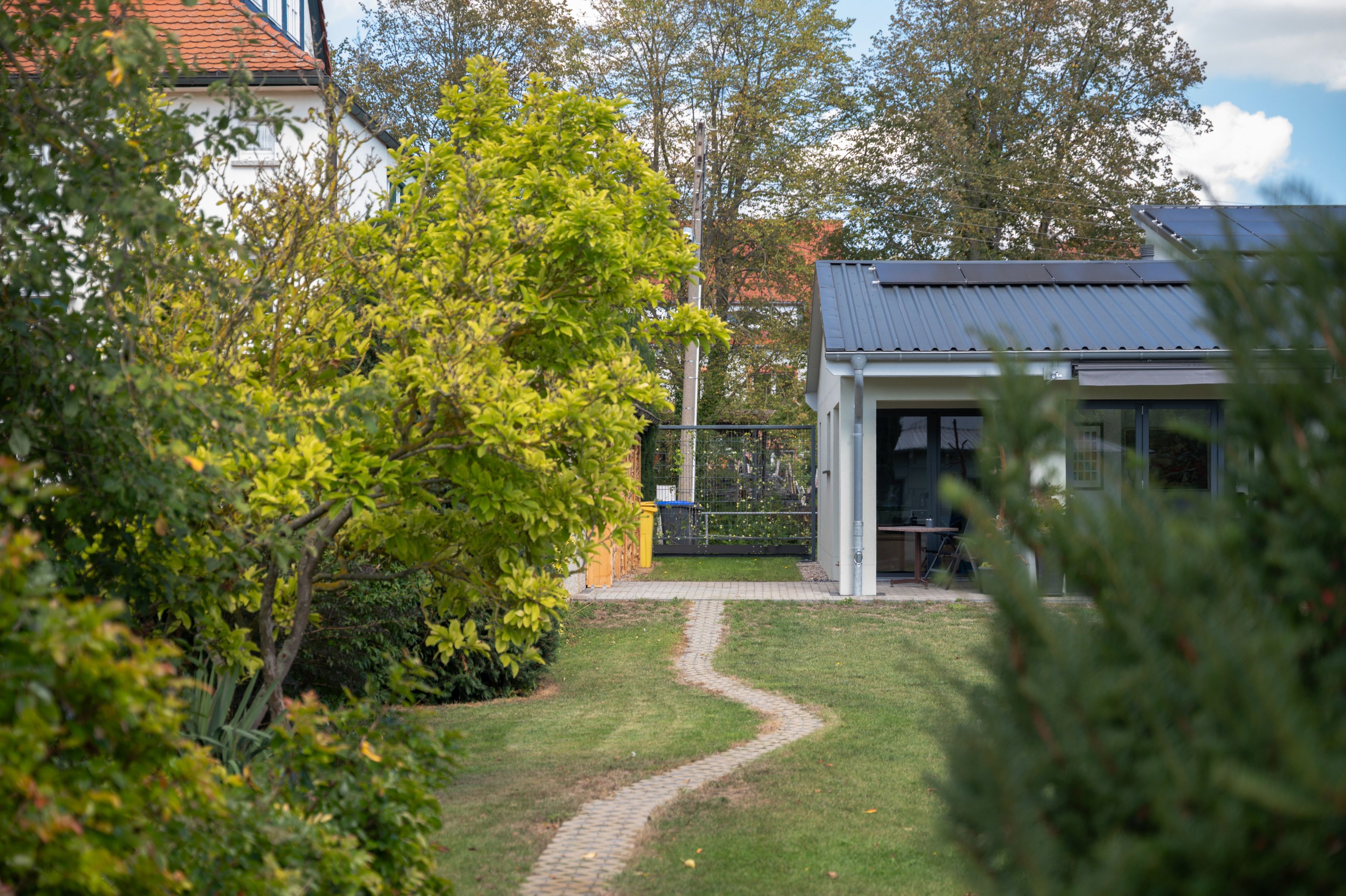 Ein Weg führt zu einem Gebäude mit Blechdach, umgeben von Grünflächen und Bäumen.
