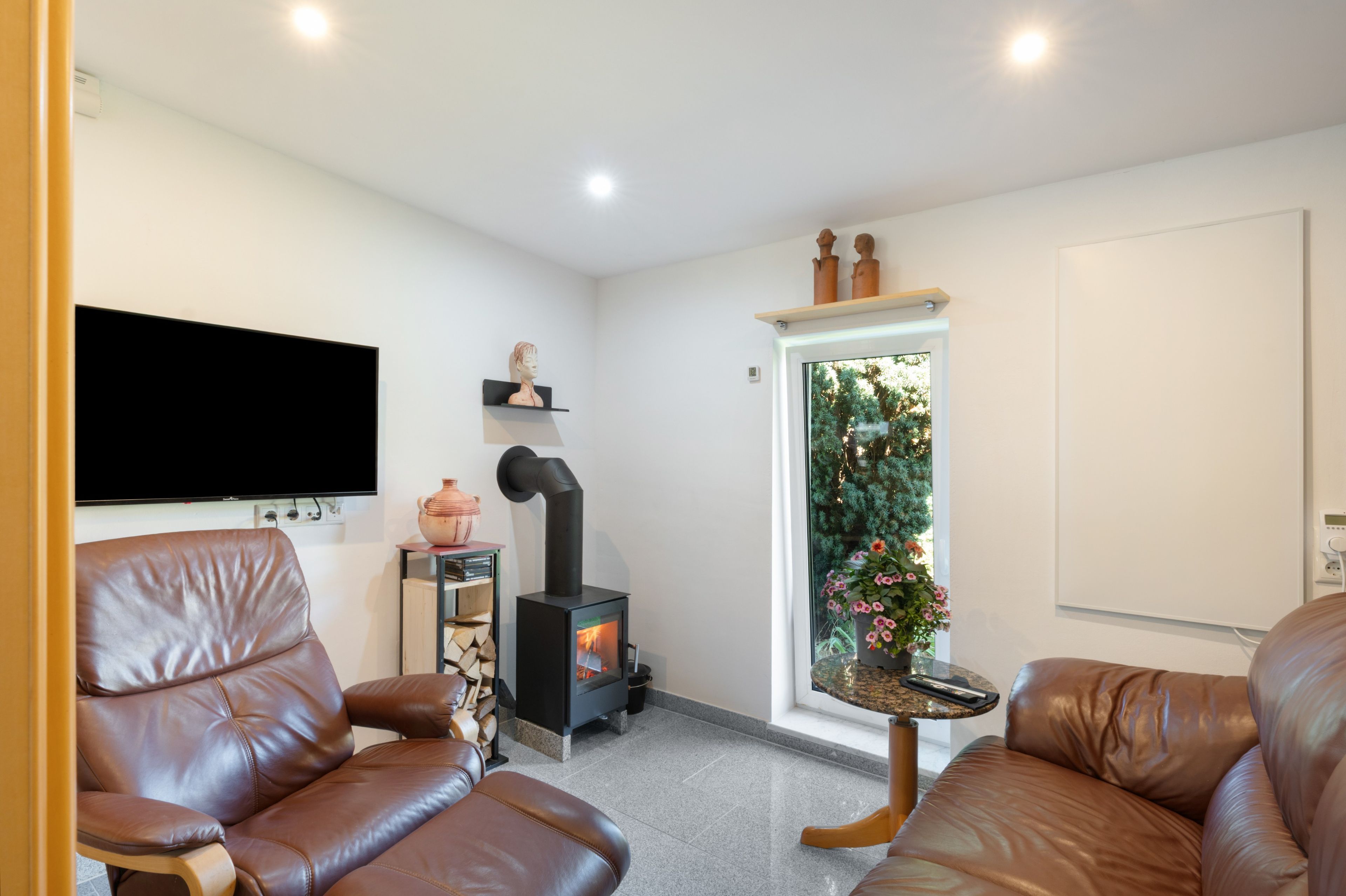 A modern living room with leather sofas, a flat-screen TV, a wood-burning stove, and a window with a view of greenery.