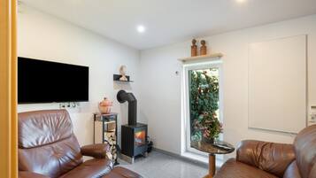 A modern living room with leather sofas, a flat-screen TV, a wood-burning stove, and a window with a view of greenery.