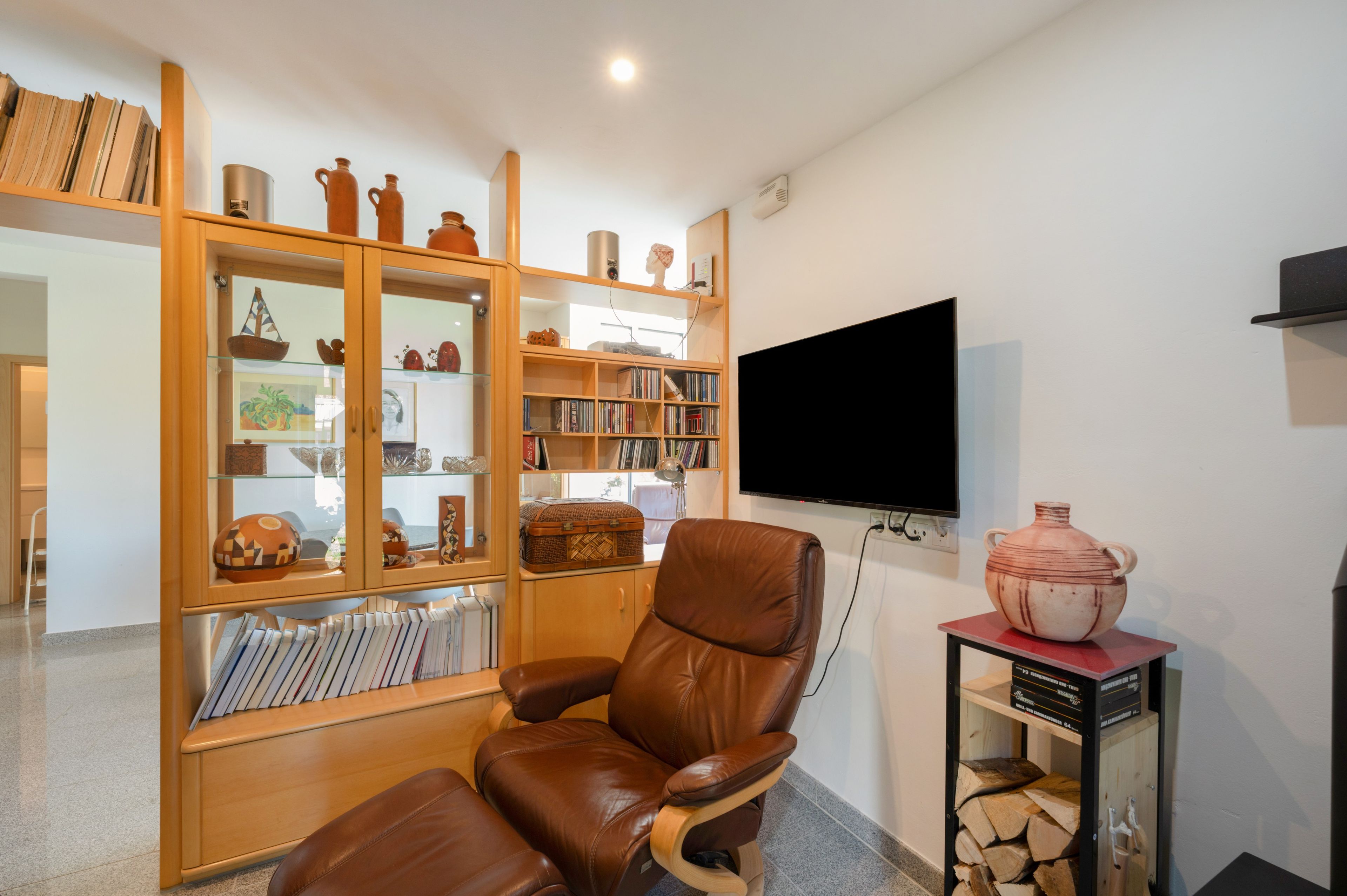 Sala de estar com poltrona de couro, estante repleta de livros e objetos decorativos, televisão de tela plana na parede e lareira com espaço para lenha embaixo.