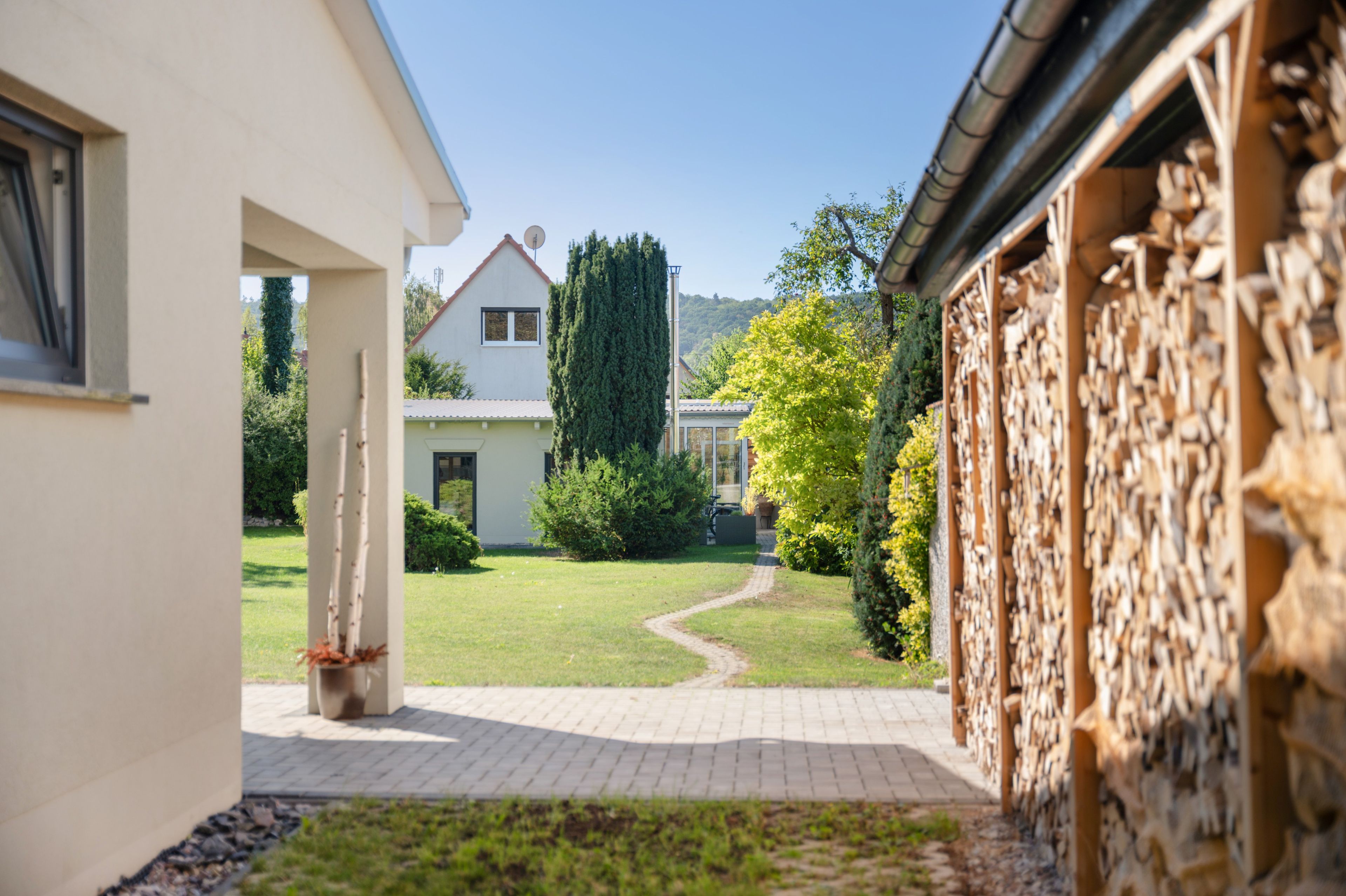 Ein Weg führt zu einem Haus mit weißer Fassade, einem Garten mit Rasen und Bäumen sowie einer Steinmauer auf der rechten Seite.