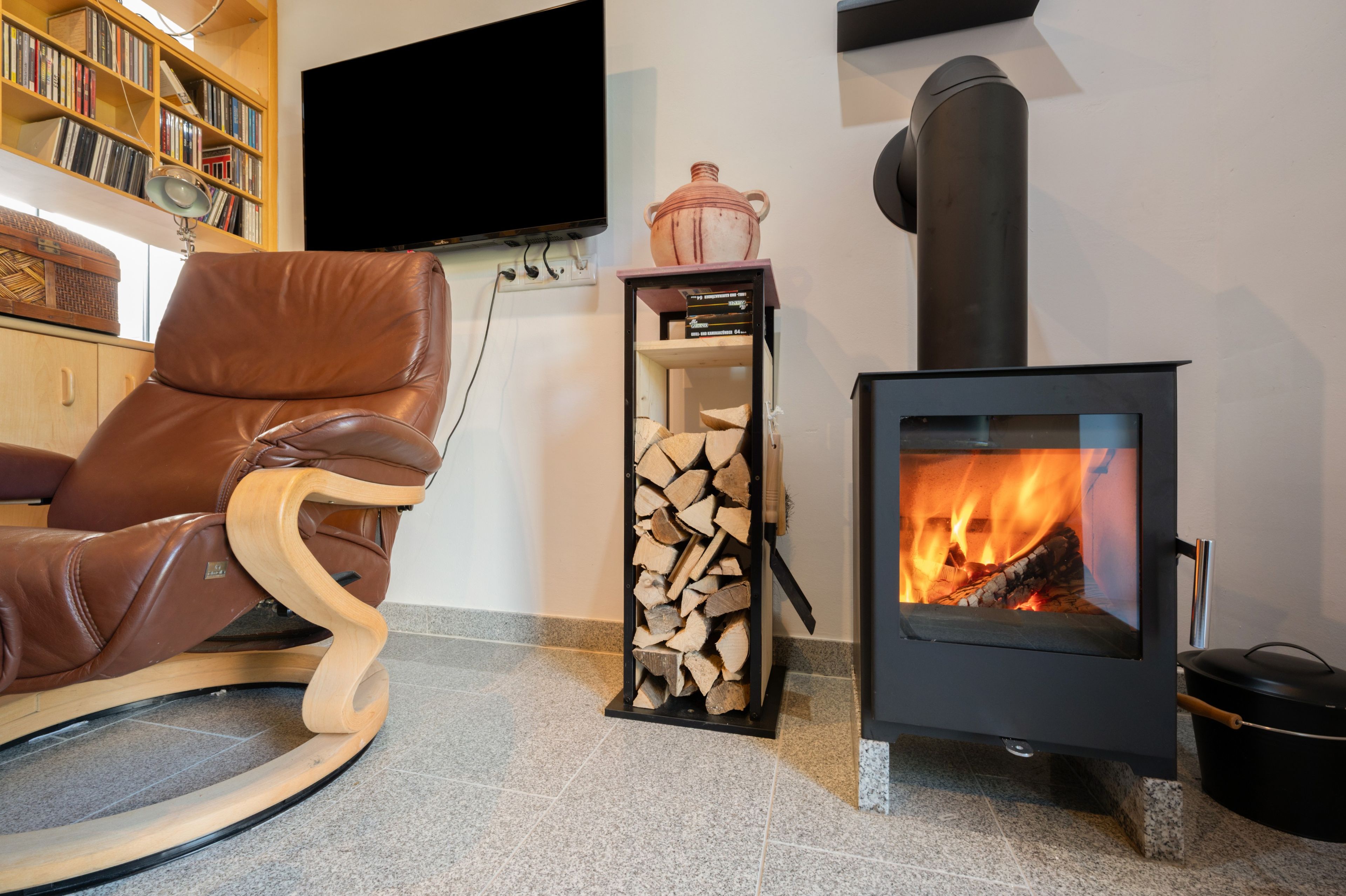 A cozy living room with a leather armchair, a wood-burning stove, a bookshelf, and a flat-screen TV.