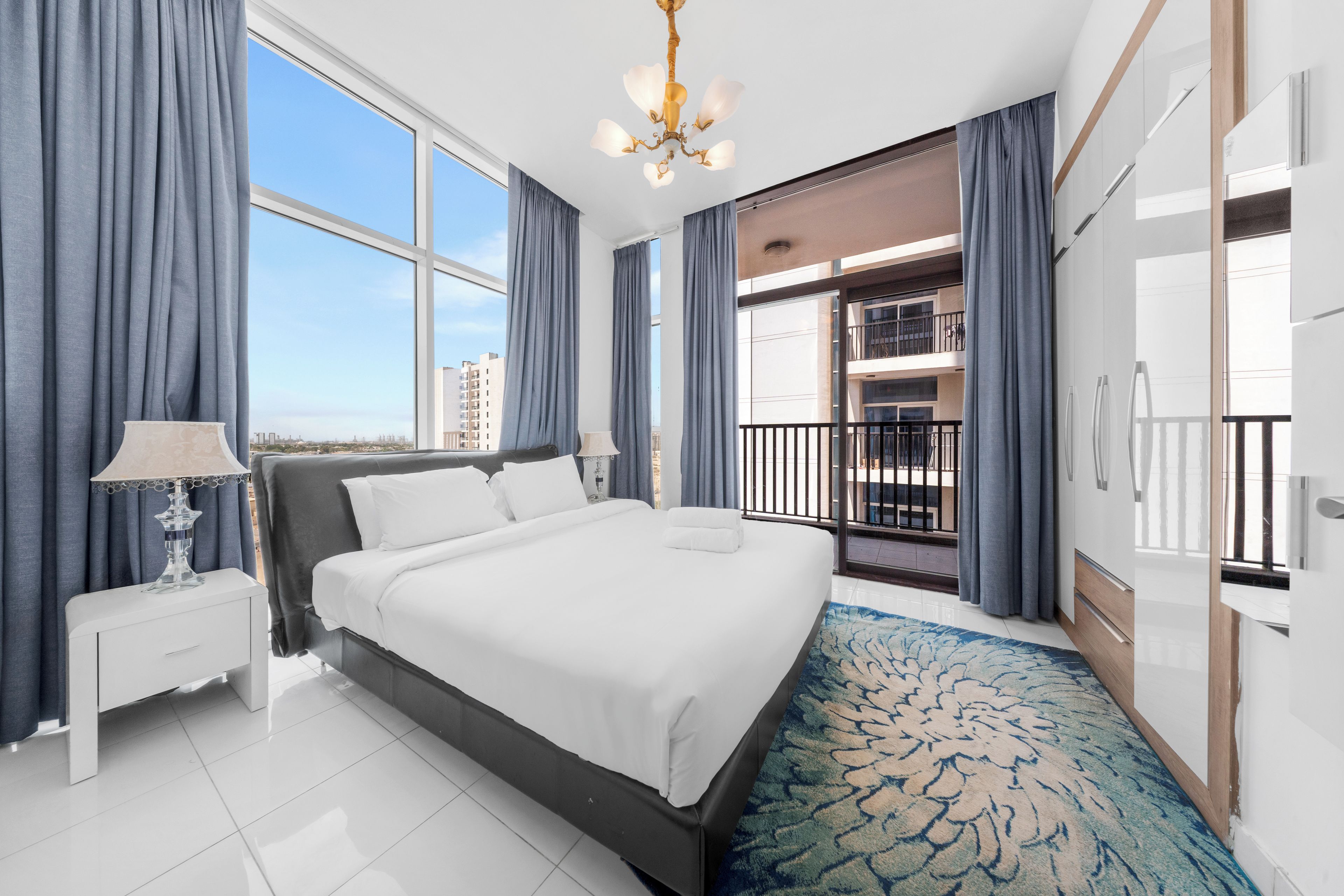 A hotel room with a large bed, a nightstand, a lamp, a view of the cityscape, and a balcony with a railing.