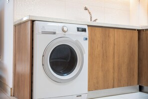 A front-loading washing machine in a kitchen with wooden cabinets and a white countertop. - StayShort - Peaceful 2-bedroom in Al Furjan that Sleeps 6! (Al Furjan)