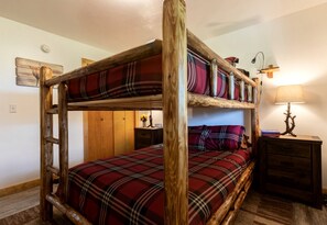 Una cama litera con ropa de cama a cuadros, una escalera de madera, una mesita de noche con una lámpara y un estante fijado a la pared.