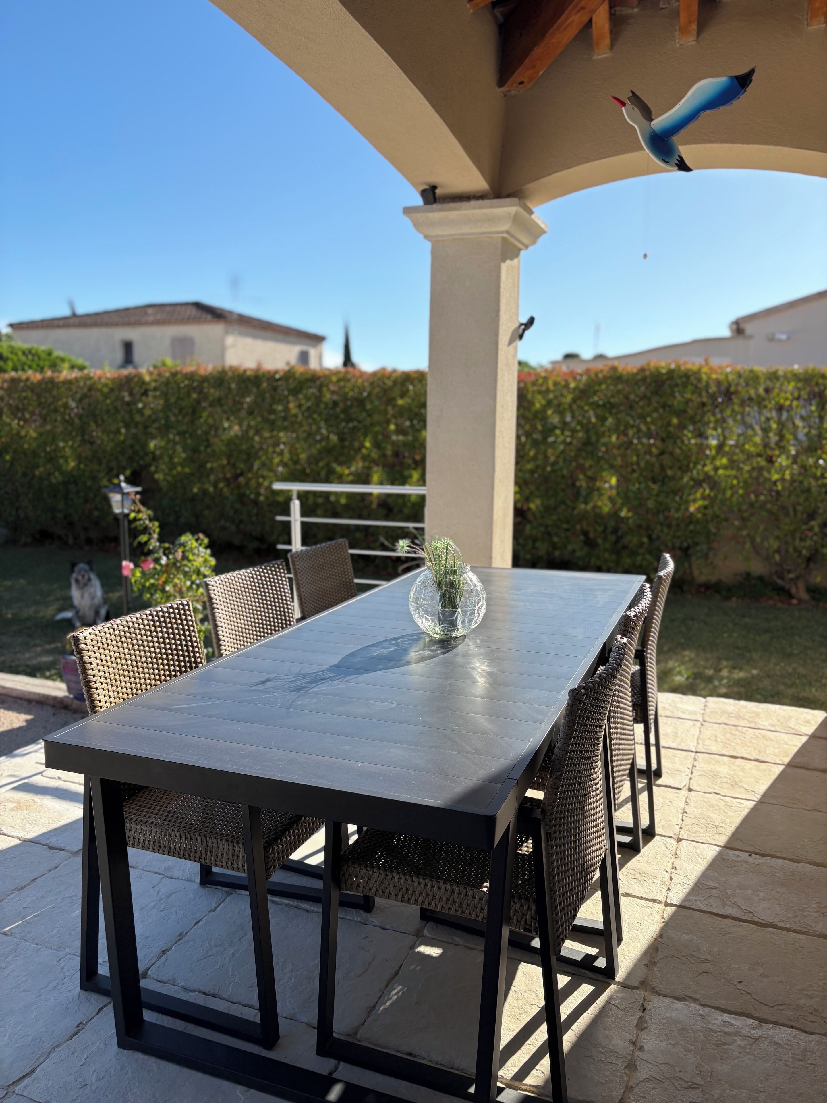 An outdoor patio with a rectangular table and wicker chairs, a decorative vase, and a bird sculpture.