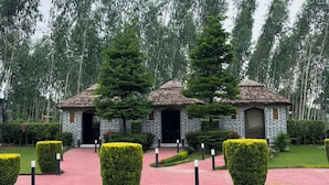 A building with a brick exterior and a thatched roof, surrounded by well-manicured hedges and a paved walkway. - Farmgrill Restaurant And Party Lawn (Haldwani)