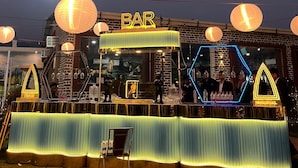 A bar with a neon sign, illuminated by hanging lanterns, and a hexagonal structure with people inside. - Farmgrill Restaurant And Party Lawn (Haldwani)