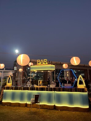 Un bar con un letrero de neón, iluminado por faroles colgantes, y una estructura hexagonal con gente en su interior.