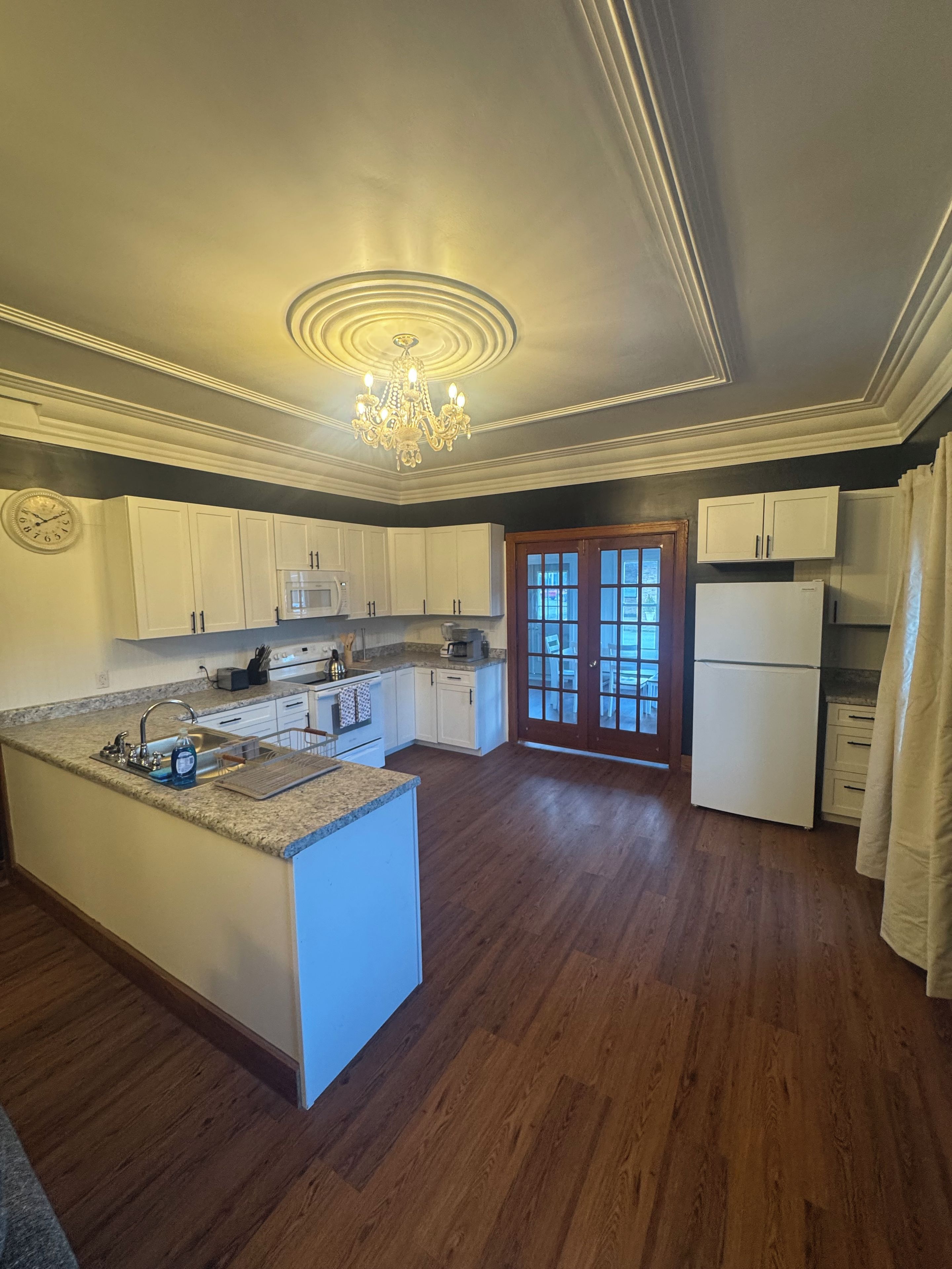 Una cocina amplia con una isla central, muebles blancos, una araña y un par de puertas que dan a un espacio exterior.