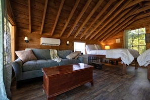 A cozy cabin with a wooden ceiling, a sofa, a wooden coffee table, a bed, and a window with a view of trees.