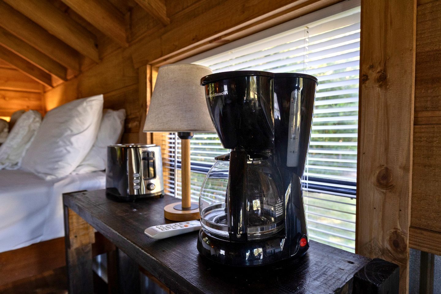 A wooden cabin interior with a bed, a lamp, a coffee maker, and a remote control on a wooden table.