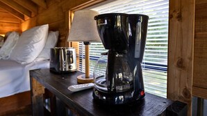Interieur van een houten hutje met een bed, een lamp, een koffiezetapparaat en een afstandsbediening op een houten tafel.