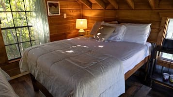 Quarto de cabana de madeira com cama, mesa de cabeceira, abajur e janela com vista para árvores.