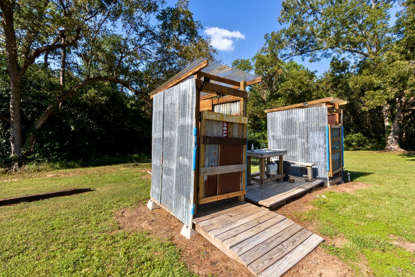 Dos pequeñas construcciones rústicas de madera con techos de metal, cada una con una terraza de madera que conduce a un banco y una mesa, ubicadas en un área con césped y árboles al fondo.