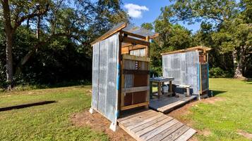 Dos pequeñas construcciones rústicas de madera con techos de metal, cada una con una terraza de madera que conduce a un banco y una mesa, ubicadas en un área con césped y árboles al fondo.