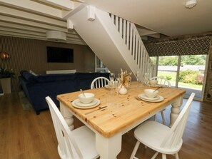 A dining area with a wooden table set for four, white chairs, a staircase, a sofa, and a television in the background. - The Cart House (Llandysul)
