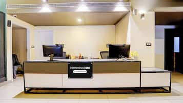 Un mostrador de recepción con un cartel que indica servicio de empresa. Hay dos monitores de computadora, un teclado y un ratón en el mostrador. Una silla se encuentra a la izquierda del mostrador.