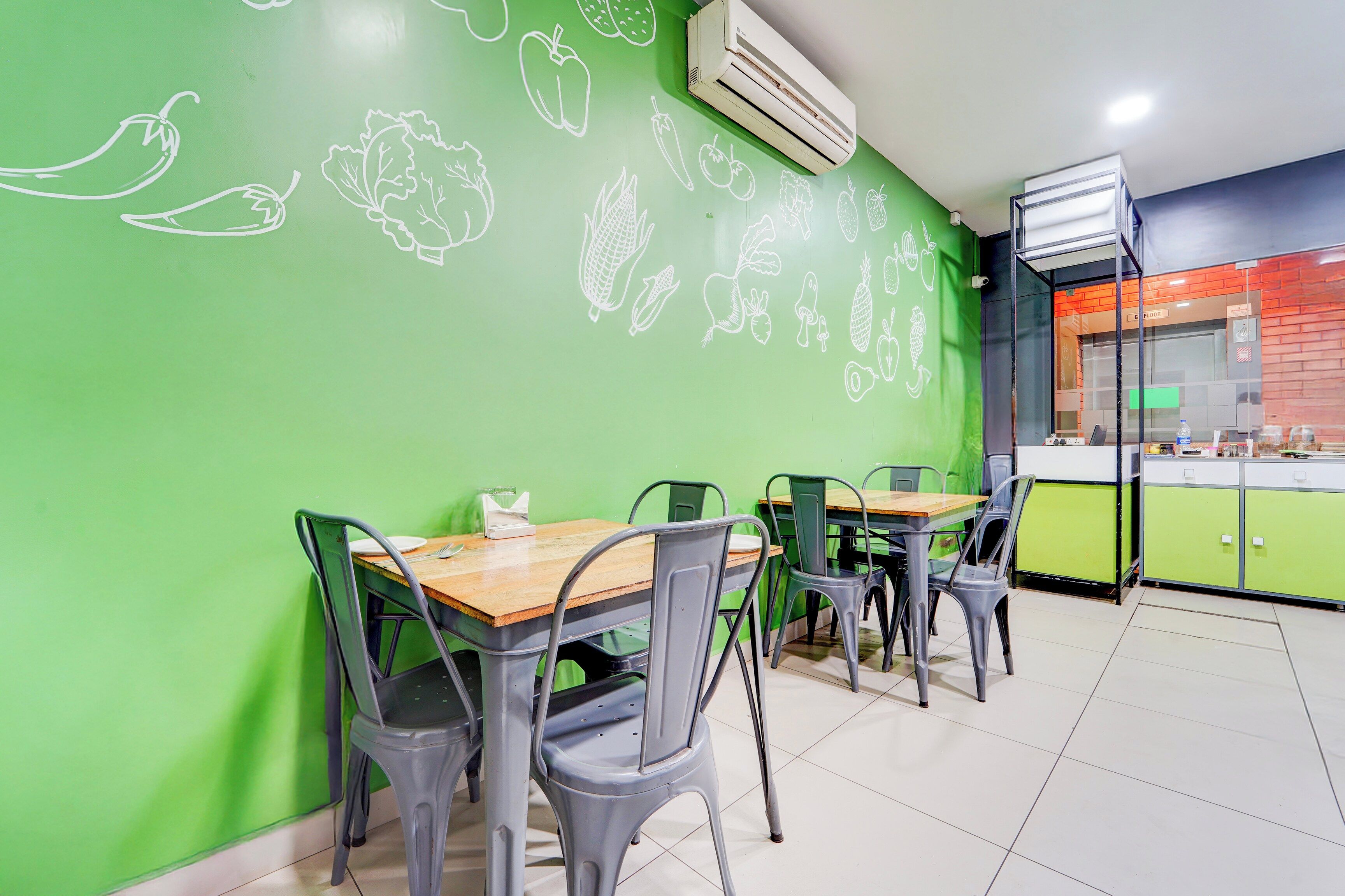 A restaurant interior with green walls, whiteboard drawings of food items, and metal chairs around tables.