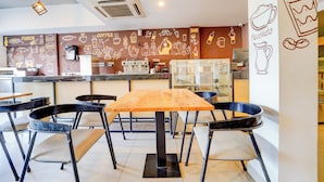 Interior de una cafetería con una mesa de madera, sillas negras, una máquina de café y obras de arte que representan elementos relacionados con el café.