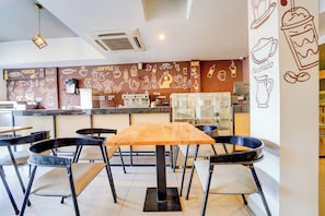 Interior de una cafetería con una mesa de madera, sillas negras, una máquina de café y obras de arte que representan elementos relacionados con el café.