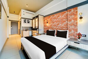 Une chambre d’hôtel moderne avec un mur d’accent en briques, un grand lit, une table de chevet agrémentée d’un vase de fleurs et un coin cuisine.