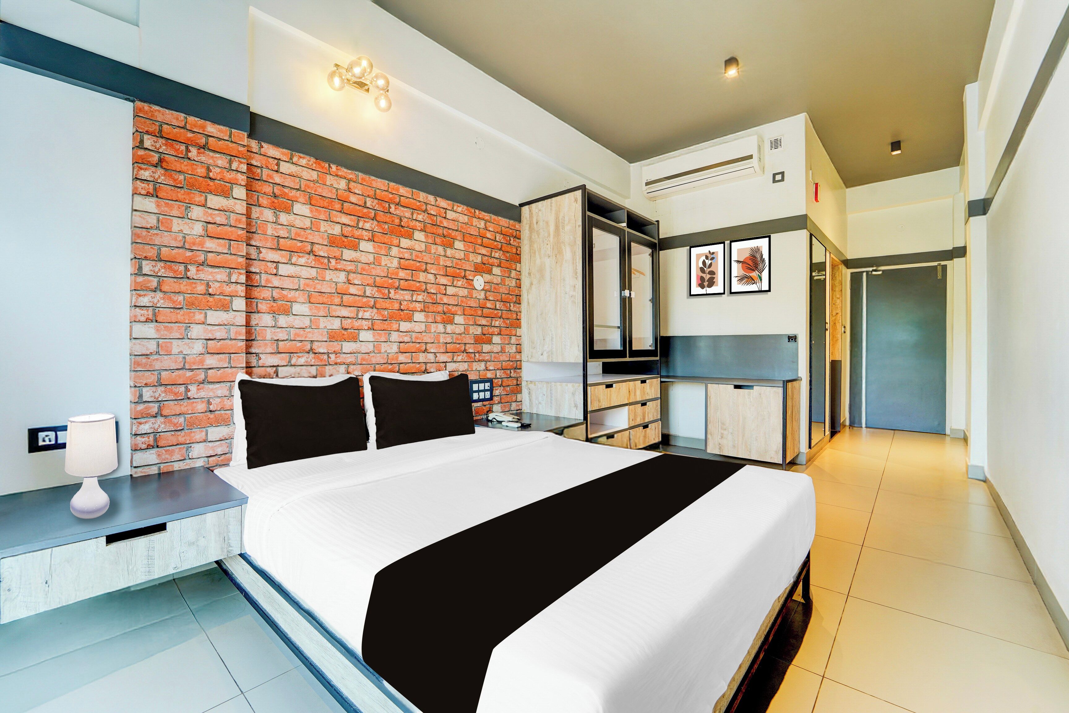 Een hotelkamer met een bakstenen muur, een bed met zwart-wit beddengoed, een nachtkastje met een lamp, een kledingkast en een kitchenette.
