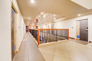 A multi-level hotel corridor with a tiled floor, a metal railing, and doors leading to rooms.
