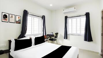 A hotel room with a bed, two windows with curtains, a desk with a chair, and an air conditioning unit.