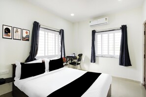 Habitación de hotel con una cama, dos ventanas con cortinas, un escritorio con una silla y un equipo de aire acondicionado.