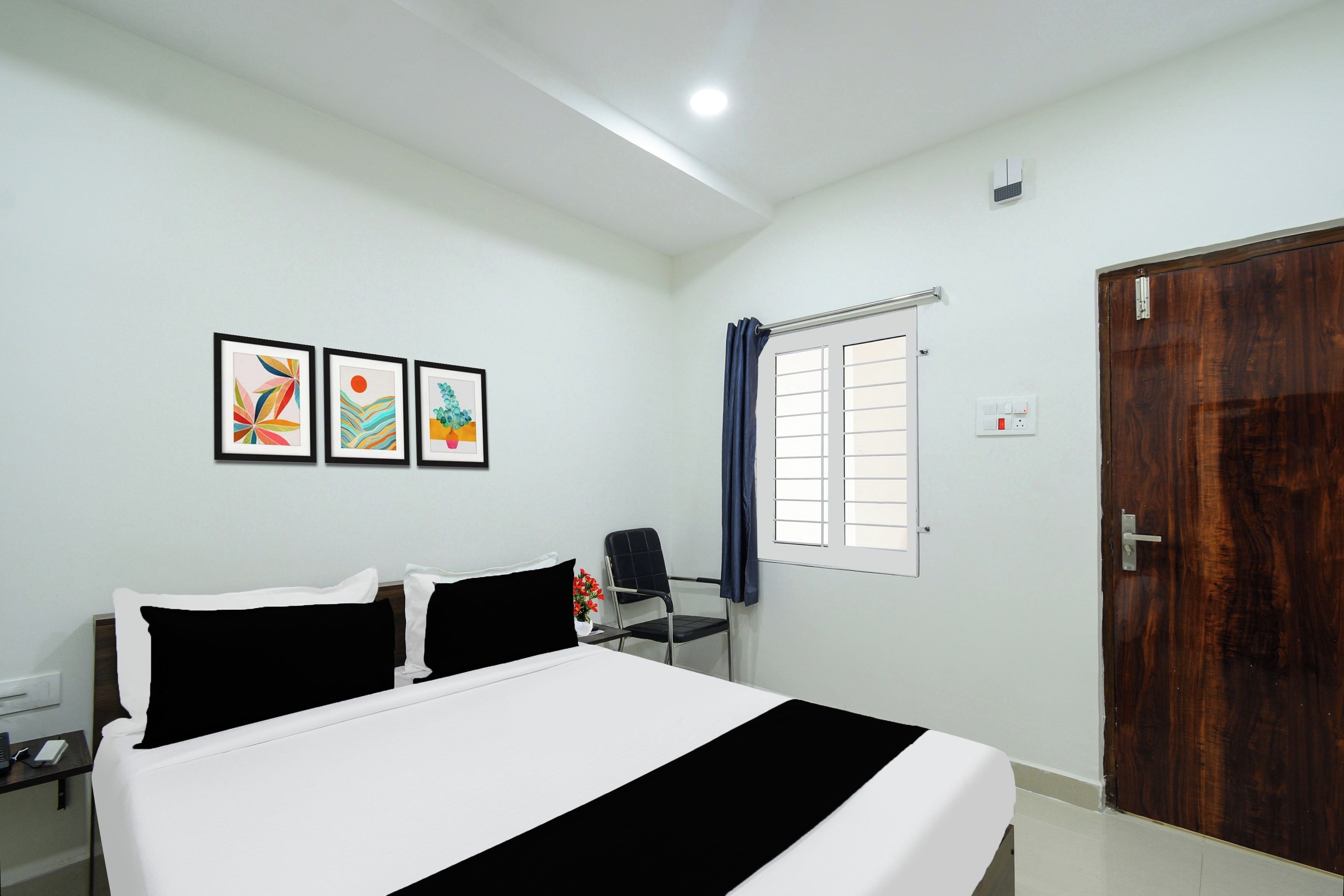 A hotel room with a bed, a desk, a chair, a window with curtains, and three framed pictures on the wall.