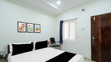 A hotel room with a bed, a desk, a chair, a window with curtains, and three framed pictures on the wall.