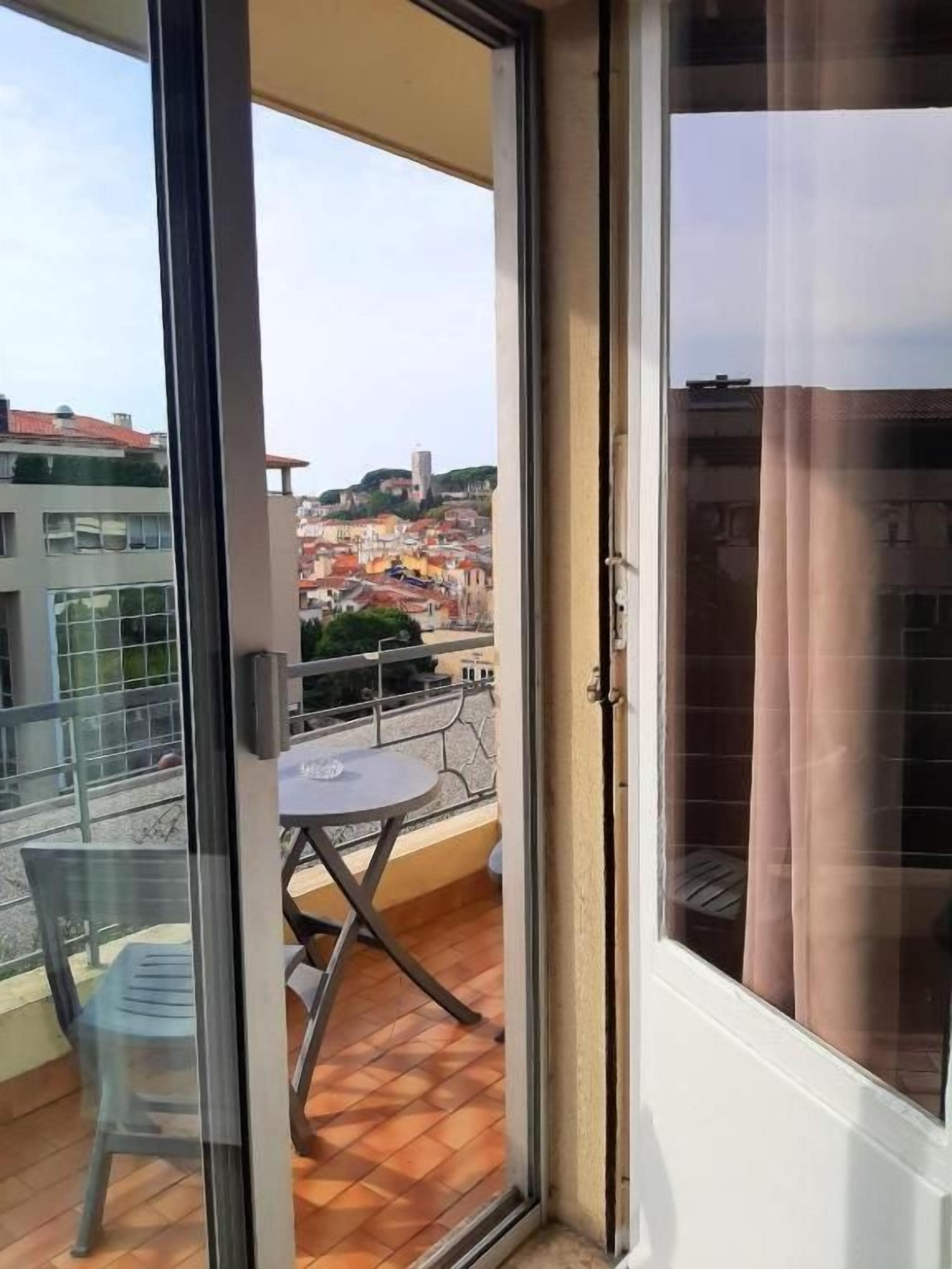 Un balcón con mesa y sillas, una puerta corredera de vidrio y vista a un paisaje urbano.