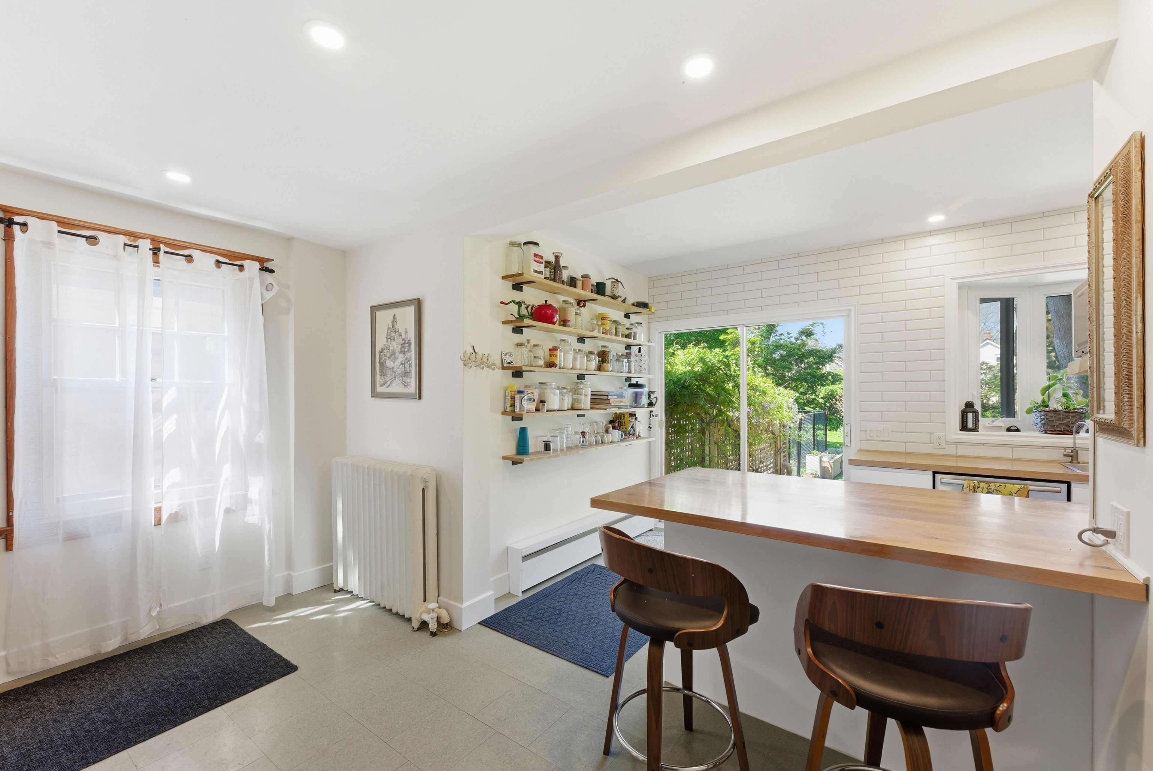 A modern kitchen with a wooden countertop, two bar stools, a radiator, and a window with a view of greenery.