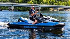 Uma pessoa andando em um jet ski azul e branco sobre a água, com uma ponte e árvores ao fundo.