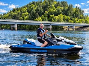 Une personne conduisant un jet ski bleu et blanc sur l’eau, avec un pont et des arbres en arrière-plan.