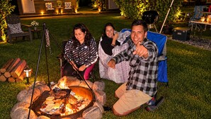 Um grupo de pessoas sentadas ao redor de uma fogueira em uma área aberta com grama, com uma casa ao fundo.