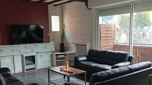 A modern living room with a fireplace, black leather sofas, a dining table, and a large window overlooking a residential area. - Lourdes: 5-Bedroom Duplex for 15 Guests, Central Location Near the Shrine (Lourdes)