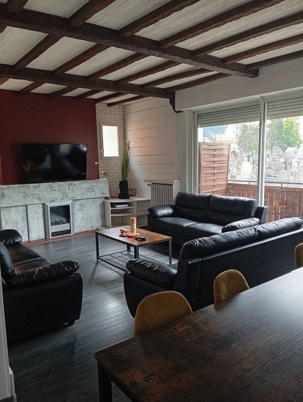 A modern living room with a fireplace, black leather sofas, a dining table, and a large window overlooking a residential area. - Lourdes: 5-Bedroom Duplex for 15 Guests, Central Location Near the Shrine (Lourdes)