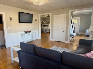 A living room with a sofa, a coffee table, and a television. There is a door leading to a kitchen and a dining area. - Historic 5-bedroom house in Swanton perfect for family getaways (Swanton)