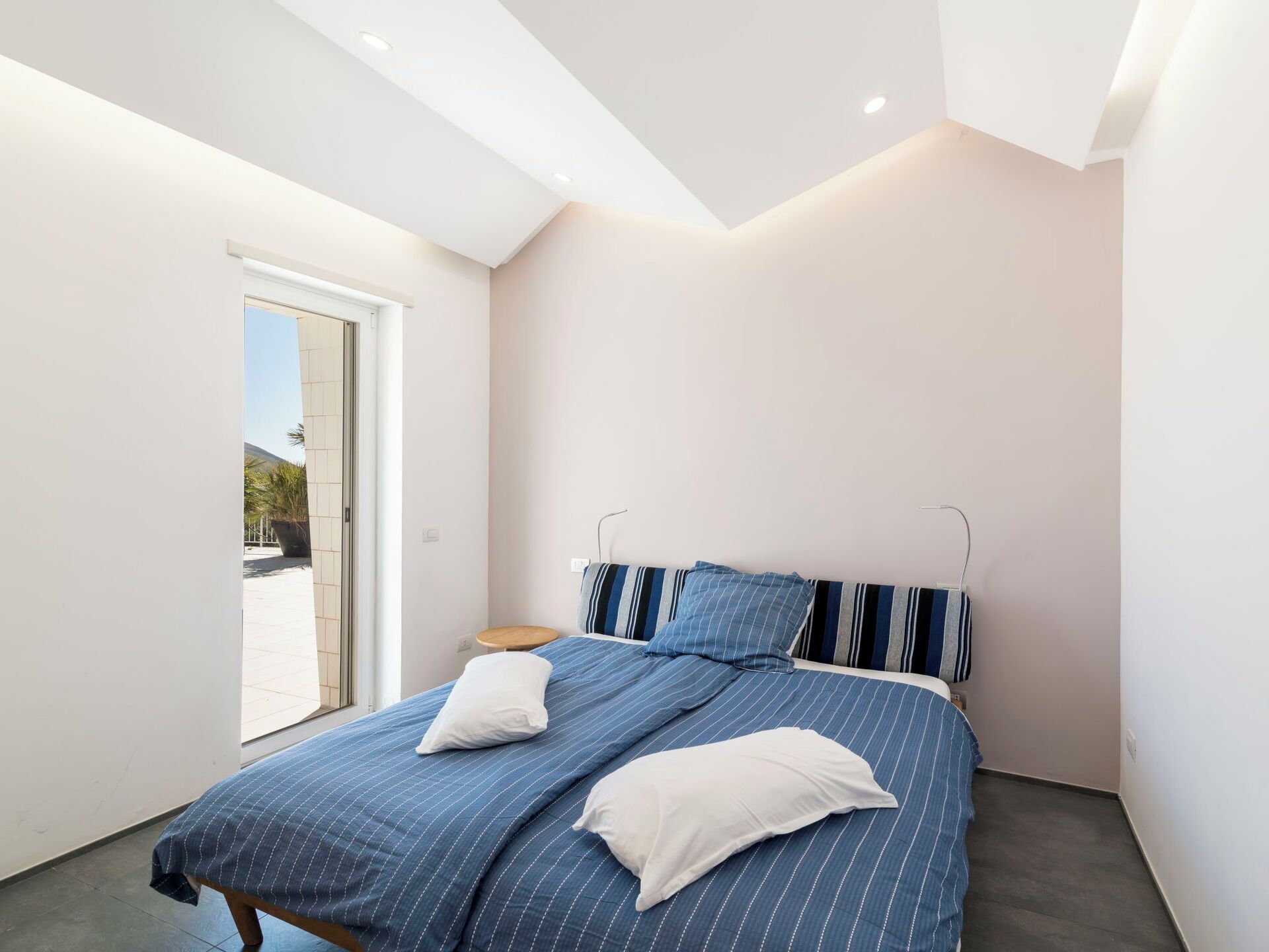 A bedroom with a bed, pillows, a bedside table, and a view through a sliding door.