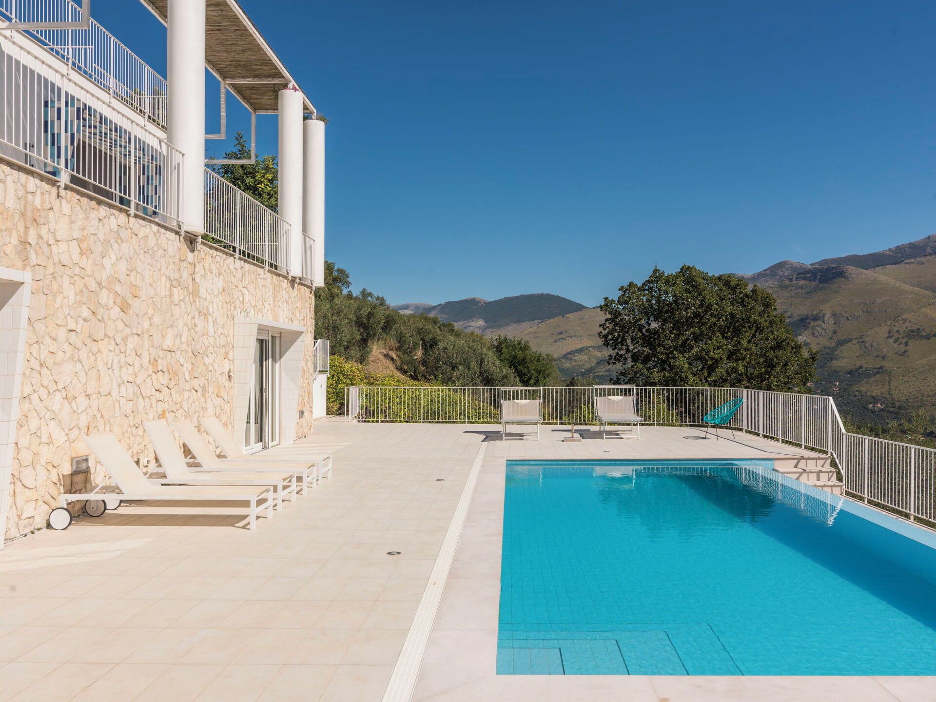 A pool area with a stone wall, lounge chairs, and a view of mountains.