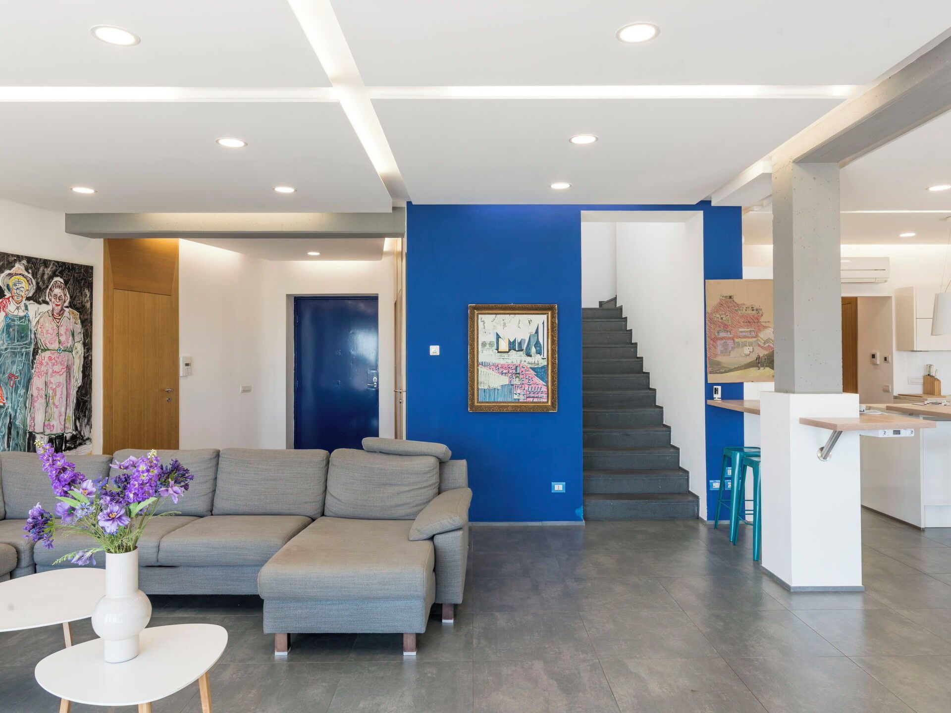 A modern living room with a grey sectional sofa, a white coffee table, and a vase with purple flowers. There is a staircase leading to a kitchen area with a blue accent wall and a framed painting.