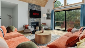 A modern living room with a stone fireplace, a flat-screen TV mounted on the wall, and a large sliding glass door leading to an outdoor deck. - Close to Sunday River Hot Tub Sauna Cold Plunge (Bethel)