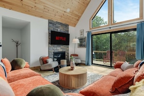 A modern living room with a stone fireplace, a flat-screen TV mounted on the wall, and a large sliding glass door leading to an outdoor deck. - Close to Sunday River Hot Tub Sauna Cold Plunge (Bethel)